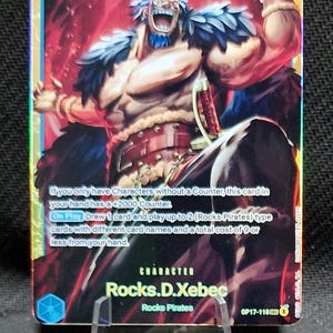 May include: A One Piece trading card featuring Rocks D. Xebec with a cost of 10 and 12000 power. The character has blue hair, a muscular build, and wears a dark cape. The card text details game mechanics, including a +2000 counter and On Play ability.