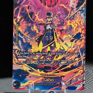 May include: A Sabo character card from the One Piece trading card game. The card features Sabo in a black coat and hat, with a fiery background. The card has a counter value of +2000 and a cost of 6.