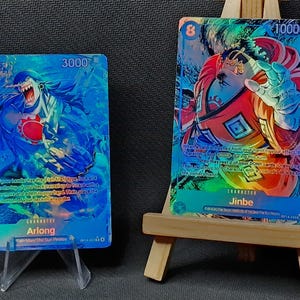 May include: Two collectible trading cards. One card features Arlong in blue, the other Jinbe in red and orange. Both cards have holographic foil effects and are displayed on small wooden easels.