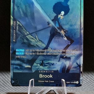 Könnte beinhalten: Eine blaue und silberne holografische Sammelkarte mit einem Charakter aus dem Anime One Piece. Der Charakter ist Brook, ein Mitglied der Strohhut-Bande. Die Karte hat eine Stärke von 6000 und einen Zählerwert von +1000. Der Kartentext lautet: "Beim Ausspielen füge bis zu 1 [Laboon] von deinem Ablagestapel zu deiner Hand hinzu. Beim K.O. spiele bis zu 1 [Laboon] mit Kosten von 4 oder weniger von deiner Hand."