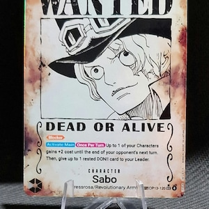 May include: A trading card featuring Sabo from the anime series. The card has a black and white illustration of Sabo with the word "WANTED" at the top. The text "DEAD OR ALIVE" is also visible. The card is held by a clear plastic stand.
