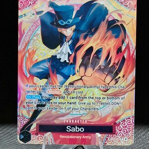 May include: A trading card featuring Sabo from the Revolutionary Army, with a dynamic illustration of the character surrounded by flames. The card has a pink and gold background with text describing its abilities. The card is displayed on a clear plastic stand.
