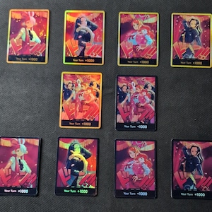Peut inclure: Dix cartes à collectionner avec des illustrations de style anime. Chaque carte présente un personnage différent dans des tons de rouge, rose et bleu. Le texte "Your Turn +1000" est imprimé en bas de chaque carte.
