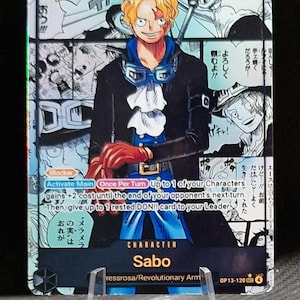Puede incluir: Una tarjeta coleccionable con el personaje Sabo de la serie One Piece. La tarjeta tiene un esquema de color azul, negro y blanco, con texto japonés y una ilustración del personaje. La tarjeta se muestra en un soporte de plástico transparente.