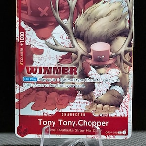 May include: A red and white trading card featuring Tony Tony. Chopper, a character with antlers and a pink hat. The card displays the number 3, the word "WINNER", and the text "COUNTER +1000". The card is propped up on a clear plastic stand.