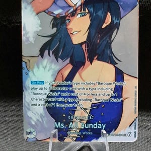 Può includere: Una carta collezionabile con un'illustrazione di Ms. All Sunday con capelli scuri e un abito blu. La carta mostra un livello di potenza di 8000, un costo di 7 e un testo che descrive le meccaniche di gioco per le carte personaggio Baroque Works.