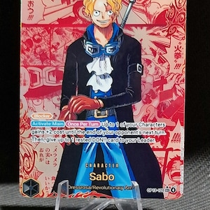 May include: A trading card featuring the character Sabo from the anime series. The card has a red background with Japanese text and illustrations. Sabo is depicted in a black coat, blue shirt, and goggles, holding a staff. The card is displayed on a clear plastic stand.