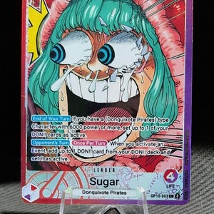 Könnte beinhalten: Sammelkarte mit Sugar von den Donquixote Pirates. Die Karte zeigt eine farbenfrohe Illustration eines Charakters mit grünen Haaren und großen Augen. Die Karte wird auf einem transparenten Kunststoffständer präsentiert.