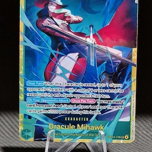 May include: A trading card featuring Dracule Mihawk from the "One Piece" series. The card has a gold and blue color scheme with an illustration of a character holding a sword. The card's text includes the character's name, game rules, and a power level of 10000.