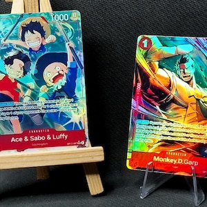 May include: Two colorful trading cards featuring anime characters. One card displays Ace, Sabo, and Luffy with the text "Ace & Sabo & Luffy". The other card shows Monkey D. Garp with the text "Monkey.D.Garp". Both cards have a value of 1.