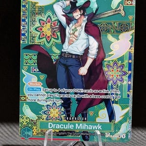 May include: Trading card featuring Dracule Mihawk from the One Piece series. The card has a teal and gold background with an illustration of the character. The card text includes "Blocker" and "On Play" instructions. The card is displayed on a clear stand.