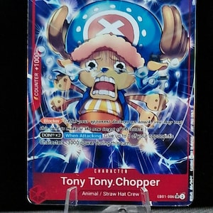 May include: A trading card featuring Tony Tony Chopper from the One Piece anime. The card has a blue background with a distressed Chopper illustration. The card text includes "Blocker" and "When Attacking". The card is displayed on a clear plastic stand.
