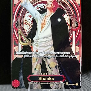 Può includere: Una carta collezionabile con il personaggio Shanks della serie One Piece. La carta ha un bordo decorativo rosso e oro, con il personaggio in camicia bianca e cappotto nero. La carta mostra il numero 10 e il testo "Shanks".