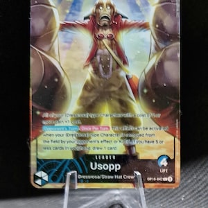 Könnte beinhalten: Eine One Piece Sammelkarte mit Usopp von der Dressrosa/Strohhut-Crew. Die Karte hat einen Wert von 5000 und enthält Spieltext. Die Figur ist mit ausgestreckten Armen dargestellt und trägt eine rote Jacke und eine beige Hose.