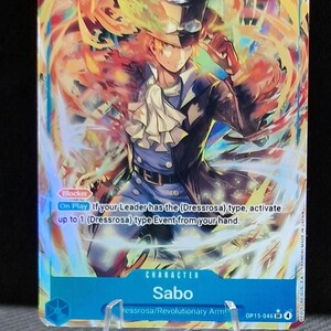May include: A collectible trading card featuring the character Sabo. The card displays Sabo in a dark blue coat, white ruffled shirt, and a top hat, set against a fiery background. The card has text that reads "Blocker" and "On Play".