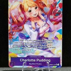 May include: A collectible trading card featuring Charlotte Pudding from the Big Mom Pirates. The card has a purple background with a character illustration, a power level of 5000, and the number 7. The card also includes game text.