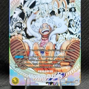 Op de afbeelding: Een One Piece ruilkaart met Monkey D. Luffy in zijn Gear 5 vorm met wit haar en kleding. De kaart bevat de tekst: If you have 1 or less Life cards, give this card in your hand -1 cost. On Play You may trash 1 card from your hand. De kaart is glanzend.