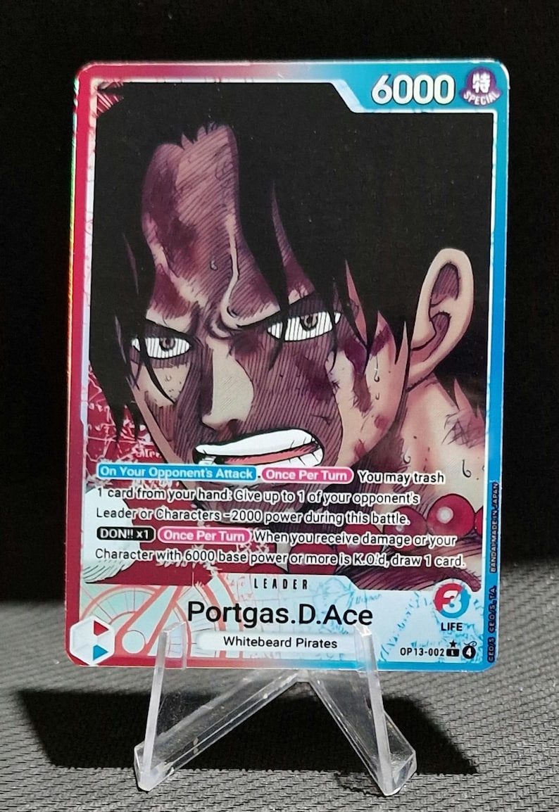 Puede incluir: Una tarjeta coleccionable con una ilustraci&oacute;n de Portgas D. Ace con una expresi&oacute;n decidida. La tarjeta tiene un borde azul y rojo con texto que indica su estado especial y sus mec&aacute;nicas de juego. La tarjeta se muestra en un soporte de pl&aacute;stico transparente.