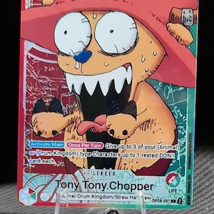 May include: A One Piece trading card featuring Tony Tony Chopper. The card has a red and green background with the character Chopper depicted with a large grin and wearing a pink hat. The card includes text such as "5000" and "STRIKE".