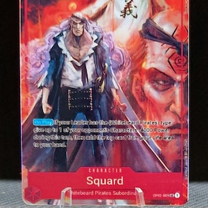 Puede incluir: Una carta de juego roja y blanca con un personaje del anime One Piece. El personaje es Squard, un subordinado de los Piratas de Barbablanca. La carta tiene un nivel de poder de 5000 y un símbolo de barra oblicua. El texto en la carta dice: "Al jugar: Si tu Líder tiene el tipo (Piratas de Barbablanca), renuncia a hasta 1 de los Personajes de tu oponente - 4000 de Poder durante este turno, luego agrega la carta superior de tu área de Vida a tu mano."