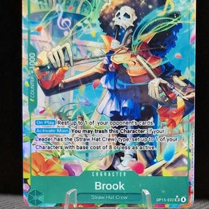Könnte beinhalten: Eine One Piece Sammelkarte mit der Figur Brook, die Geige spielt. Die Karte hat ein grünes und blaues Farbschema mit dem Text "Brook" und "Straw Hat Crew". Die Karte wird auf einem transparenten Ständer präsentiert.
