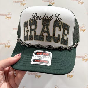 May include: A dark green and white trucker hat featuring the text Rooted In GRACE in a dark patterned font on the white foam front panel. The hat includes a silver-toned decorative chain with black oval accents across the brim base.