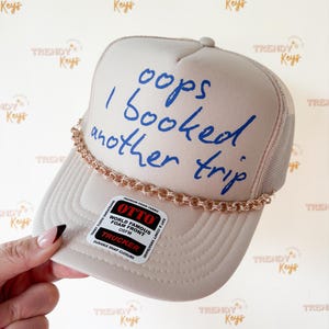 May include: A beige trucker hat featuring a foam front with the blue text "oops I booked another trip" and a metallic rose gold chain trim across the base of the crown. A rectangular sticker on the brim displays "OTTO" and "TRUCKER" branding.