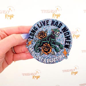 May include: A round, glittery sticker with the text "LONG LIVE RAD WOMEN". The sticker features a black panther, orange roses, and lightning bolts. The background is a mix of blue and silver glitter.
