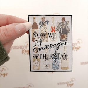 May include: A rectangular patch featuring illustrations of champagne bottles with various designs. The text "NOW WE sip champagne WHEN WE THIRSTAY" is displayed in black script. The patch is held by a hand.