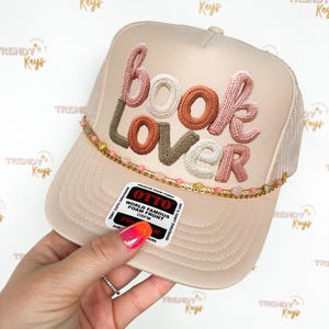 May include: A beige trucker hat with the words "BOOK LOVER" embroidered in various colors. The hat has a mesh back and a beaded accent along the brim. The hat also has a tag that says "OTTO WORLD FAMOUS FOAM FRONT OSFM".