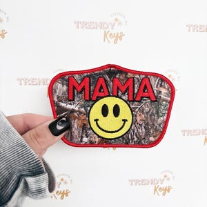 May include: Embroidered patch with the word "MAMA" in red block letters above a yellow smiley face. The patch has a camouflage background and a red border. The patch is held by a person.