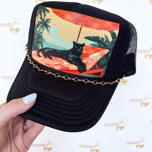 May include: Black trucker hat with a mesh back and a curved brim. The front panel features a colorful graphic of a black panther lounging on a beach chair under an umbrella. A gold chain is attached to the brim. The text "TRENDY Keys" is printed on the hat.