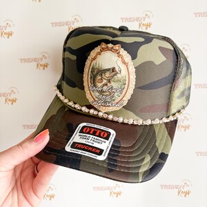 May include: Camouflage trucker hat with a decorative oval patch featuring a fish illustration and the text "hook, line, and catfished." The hat has a rhinestone-studded band and a mesh back.