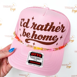 May include: Pink trucker hat with the text "I'd rather be home" in brown. The hat has a mesh back and a decorative chain with colorful links. The hat also has a "OTTO" label.