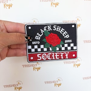 May include: Black rectangular patch with the words "BLACK SHEEP SOCIETY" embroidered in white and red. A red rose is in the center. The patch has a checkered border and the text "ESTD 1985".