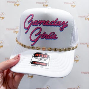 May include: A white trucker hat featuring the text Gameday Girlie in pink with a blue outline. The hat includes a gold-toned chain across the base of the front panel and a rectangular sticker on the brim that reads OTTO World Famous Foam Front.
