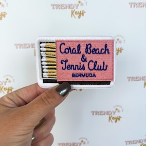 May include: Embroidered patch in the shape of a matchbook, featuring the text "Coral Beach & Tennis Club BERMUDA" in navy blue and white on a pink background. The patch includes a row of gold and black matchsticks.