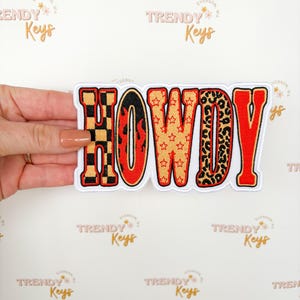 May include: A rectangular patch with the word "HOWDY" in bold, stylized letters. The letters feature various patterns: a black and red checkerboard, flames, stars, and leopard print, all outlined in red. The patch is held by a hand.