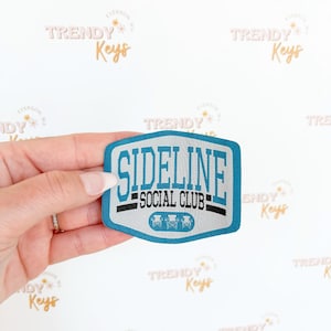Puede incluir: Un parche bordado azul y blanco con el texto "Sideline Social Club" y tres sillas estilizadas.