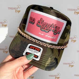 May include: A camouflage trucker hat with a pink rectangle featuring the text "no diggity, no doubt." The hat has a gold chain detail and a black mesh back. The hat is OSFM.
