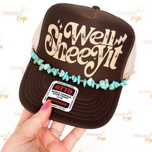 May include: A brown trucker hat with a beige mesh back and a curved brim. The hat features the text "Si Well Shee Yit" in a distressed font. A turquoise stone and gold bead trim adorns the brim. The hat has a snap closure.