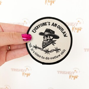 May include: Round patch with a black border and the text "EVERYONE'S AN OUTLAW" and "til it's time to do outlaw shit". The patch features a skull wearing a cowboy hat and bandana, with barbed wire.
