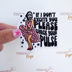 May include: Embroidered patch featuring the Ghostface mask from the Scream movies in a pink dress. The patch includes the text "IF I DON'T EXCITE YOU PLEASE CHECK YOUR PULSE" in black. The patch is outlined in white.