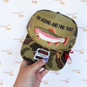 May include: A trucker hat with a camouflage pattern and a mesh back. The front of the hat features a graphic of a trout with the text "FISH AROUND AND FIND TROUT". A small charm and fishing lure are attached to the brim.