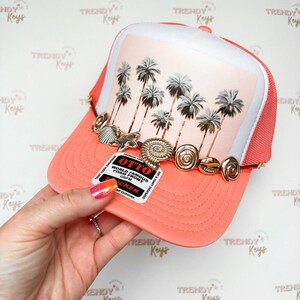 May include: A coral-colored trucker hat with a white front panel featuring a palm tree graphic. The hat is adorned with gold-colored seashell and spiral charms. The hat has a mesh back and a curved brim. The tag reads "OTTO WORLD FAMOUS FOAM FRONT OSFM".