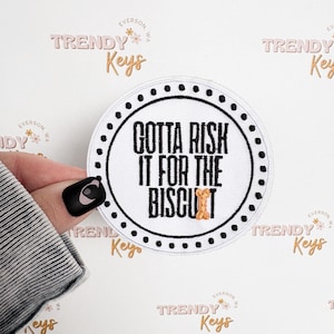 May include: A round, white patch with a black border and the text "GOTTA RISK IT FOR THE BISCUIT" in black. The patch has a small illustration of a biscuit. The background has the words "TRENDY Keys" in a repeating pattern.