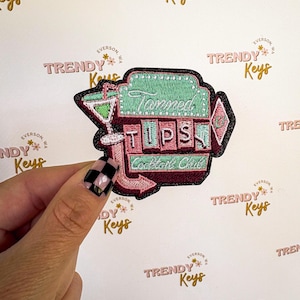 May include: Embroidered patch with a retro design. The patch features the words "Tipped Cocktail Club" in a vintage font, with a martini glass and arrow. The color scheme includes pink, green, and black.