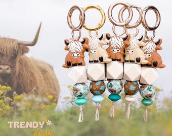 May include: Five keychains with Highland cow charms. Each keychain has a gold-colored clasp, a wooden geometric bead, and decorative beads in teal, brown, and floral patterns. The text "TRENDY Keys" is visible in the lower left corner.
