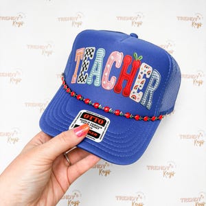May include: A blue trucker hat with the word "TEACHER" in colorful, patterned letters. The hat features a red beaded band and a mesh back. The hat is a fun accessory for teachers.