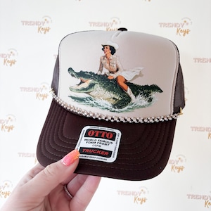 May include: Brown trucker hat with a beige front panel featuring a woman riding an alligator. The hat has a brown brim and mesh sides, embellished with rhinestones. The hat includes a "OTTO" label.